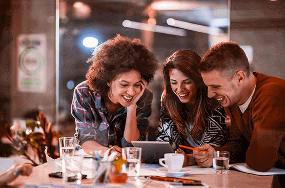 Foto de três pessoas sorrindo e olhando para um tablet, estão apoiadas em uma mesa com alguns de copos de água e lápis