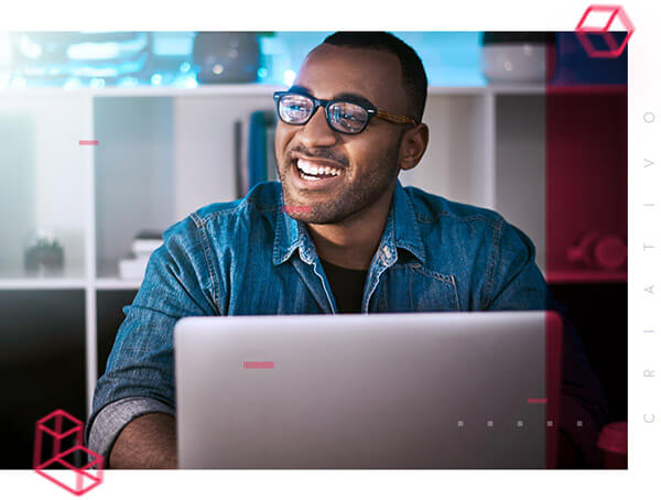 Homem moreno, cabelo curto, óculos, sorrindo, mexendo em um notebook e olhando para o lado esquerdo