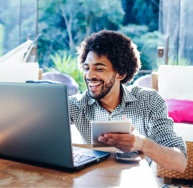 Homem sorrindo, segurando tablet; camisa xadrez; olhando para notebook; notebook em cima da mesa; natureza ao fundo